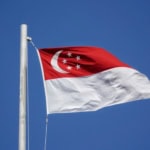 Singapore flag with red and white horizontal stripes flying on white flagpole against clear blue sky, showing crescent moon and five white stars on red field