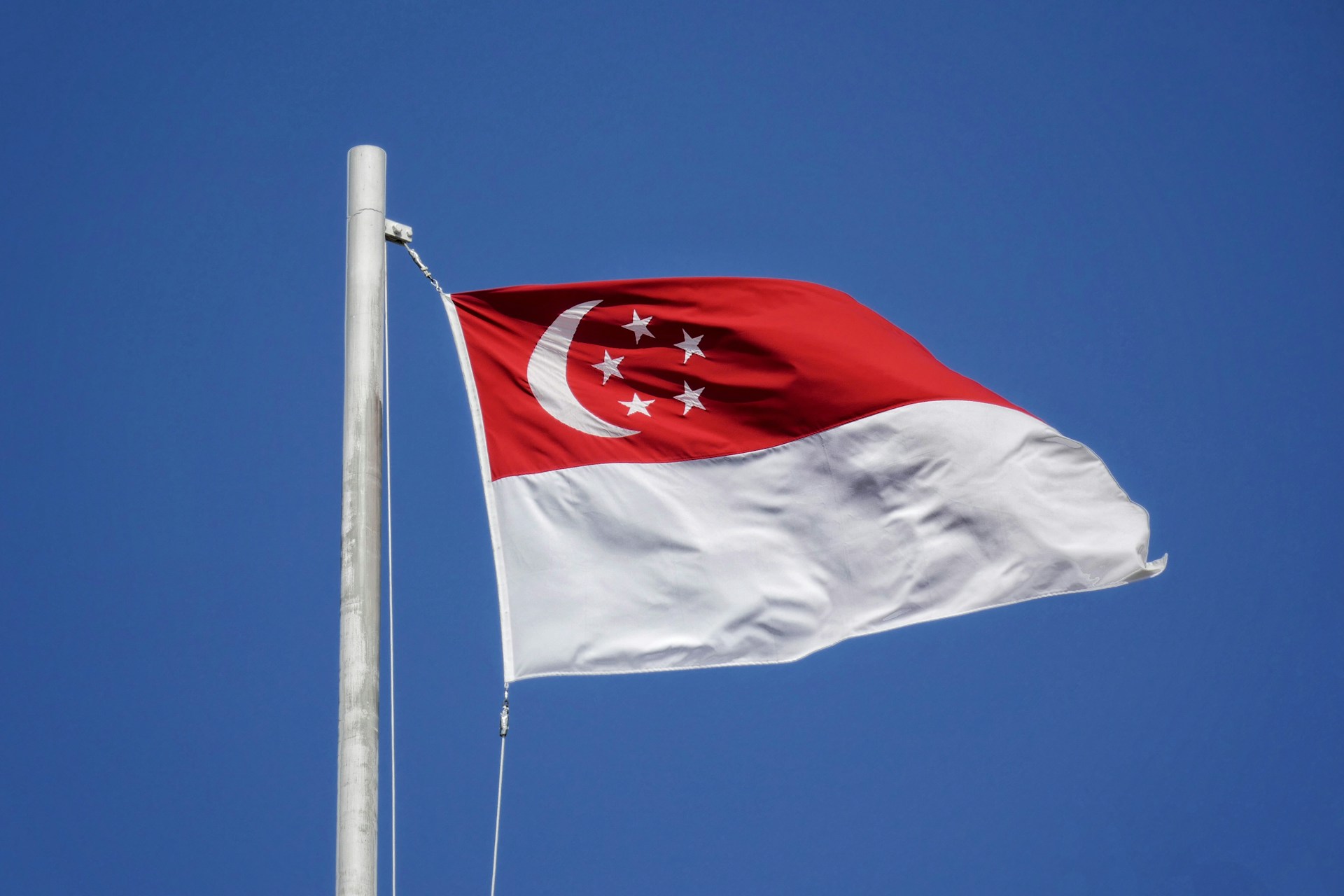 Singapore flag with red and white horizontal stripes flying on white flagpole against clear blue sky, showing crescent moon and five white stars on red field