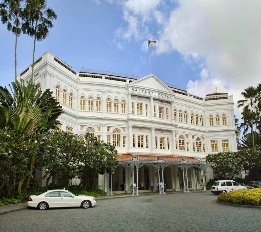 White colonial-style Raffles Hotel building with multiple stories, arched windows, and columned entrance, surrounded by palm trees with white cars in the driveway
