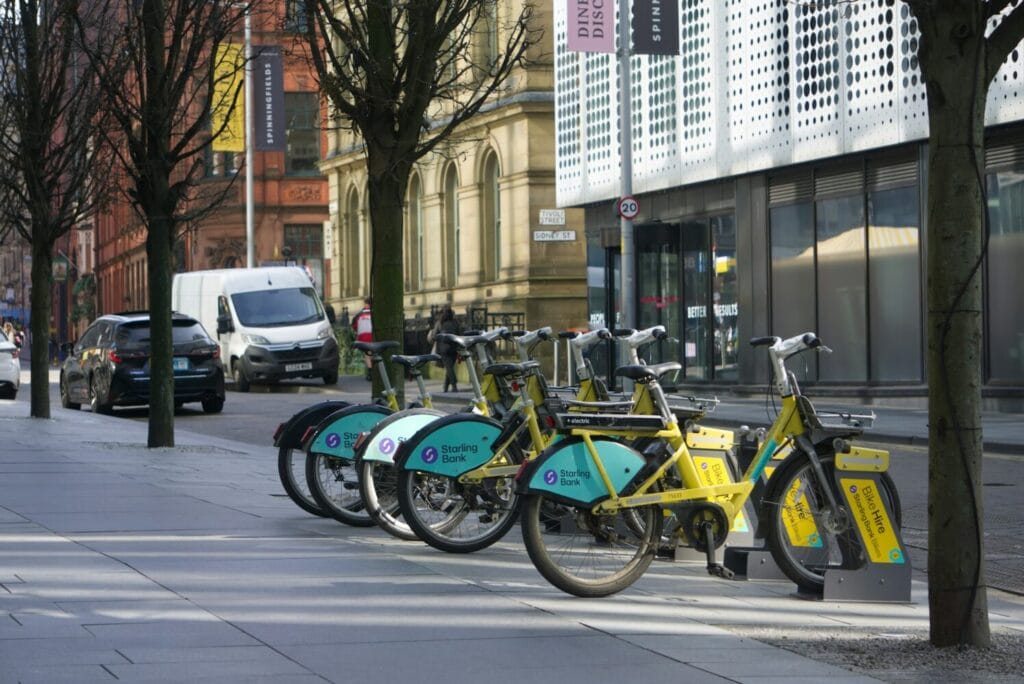 Row of yellow and teal Bee Network bike share bicycles docked at a city center station, with Sterling Bank branding visible