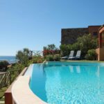 Luxury swimming pool with ocean view at Mediterranean-style villa featuring terracotta buildings and striped lounge chairs