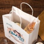 White paper bakery bag with bread and drink inside, on wooden table