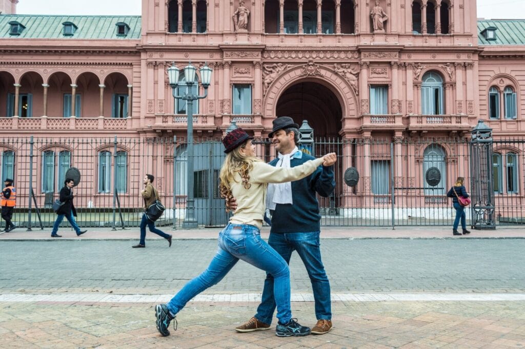 A couple dancing tango in a public square in Buenos Aires, Argentina, with a grand pink building in the background.