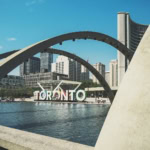 Modern concrete arches frame Toronto's downtown skyline with the iconic Toronto sign visible across a reflecting pool, featuring curved architectural elements and city skyscrapers in the background.