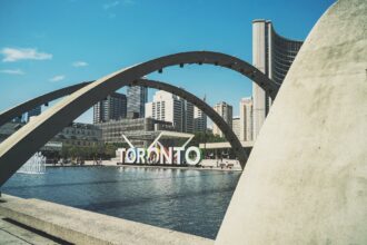 Modern concrete arches frame Toronto's downtown skyline with the iconic Toronto sign visible across a reflecting pool, featuring curved architectural elements and city skyscrapers in the background.