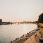 The Garonne River in Toulouse, France, is shown at sunset with people relaxing on the quayside. A bridge spans the river, and the dome of the Hôpital de La Grave is visible on the far bank.