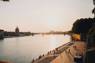 The Garonne River in Toulouse, France, is shown at sunset with people relaxing on the quayside. A bridge spans the river, and the dome of the Hôpital de La Grave is visible on the far bank.