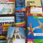 Stack of language dictionaries (Spanish, Russian, Romanian, German, Slovenian) on a desk, illustrating the need for translation apps when travelling.