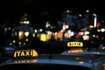 Two taxis with yellow roof signs lit up at night, parked on a blurred city street with bright lights in the background