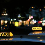 Two taxis with yellow roof signs lit up at night, parked on a blurred city street with bright lights in the background