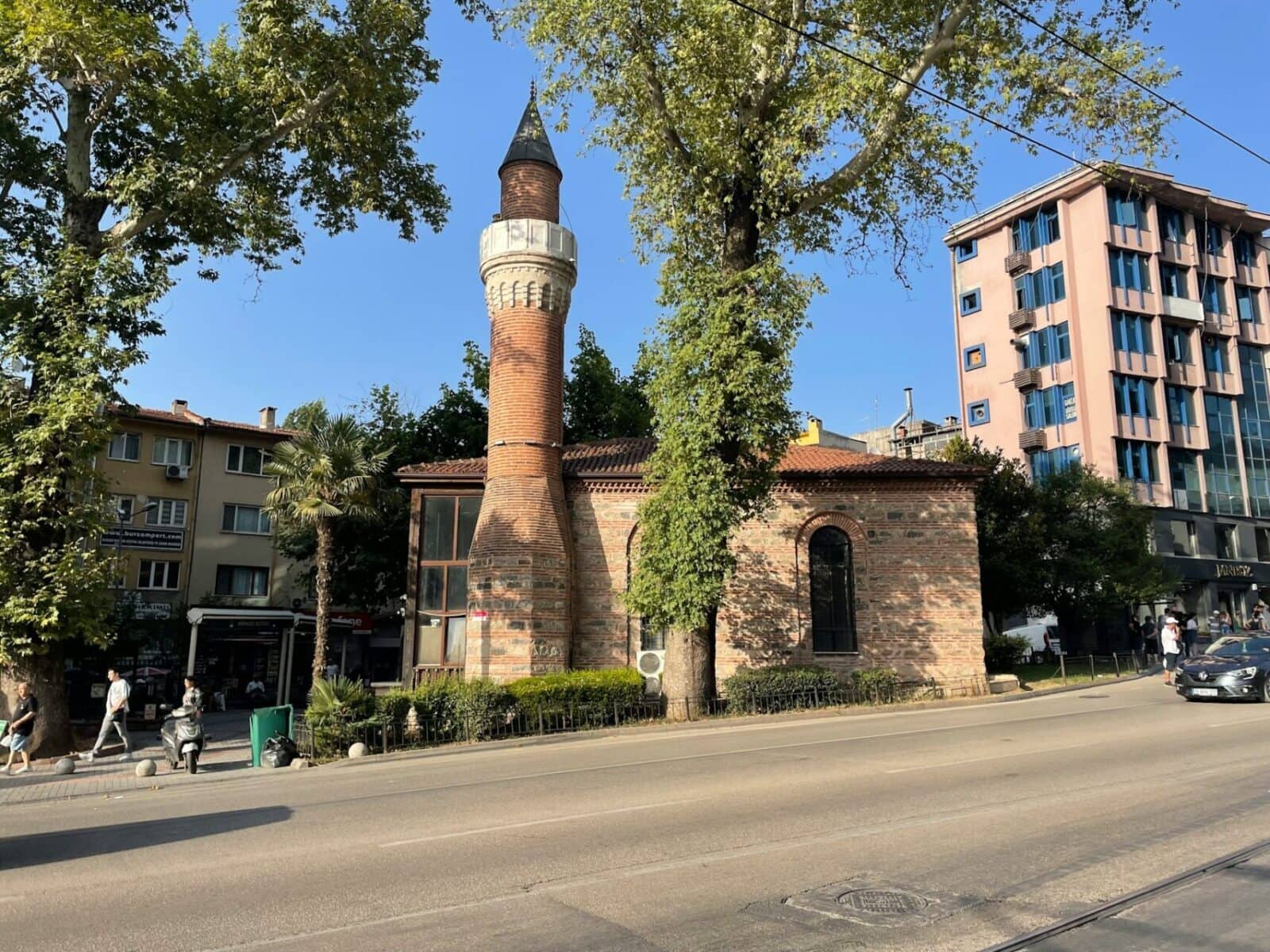 Small historic brick mosque with a tall minaret on a sunny street in Bursa, Turkey.