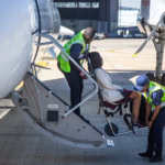 Airport staff assist an injured traveller onto an aircraft using an airport medical boarding chair. Two staff members in high-visibility vests guide the passenger safely up the aircraft steps.