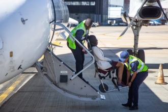 Airport staff assist an injured traveller onto an aircraft using an airport medical boarding chair. Two staff members in high-visibility vests guide the passenger safely up the aircraft steps.