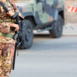 Soldier in camouflage uniform holding a rifle while standing guard beside a military vehicle at a roadside checkpoint.