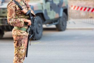 Soldier in camouflage uniform holding a rifle while standing guard beside a military vehicle at a roadside checkpoint.