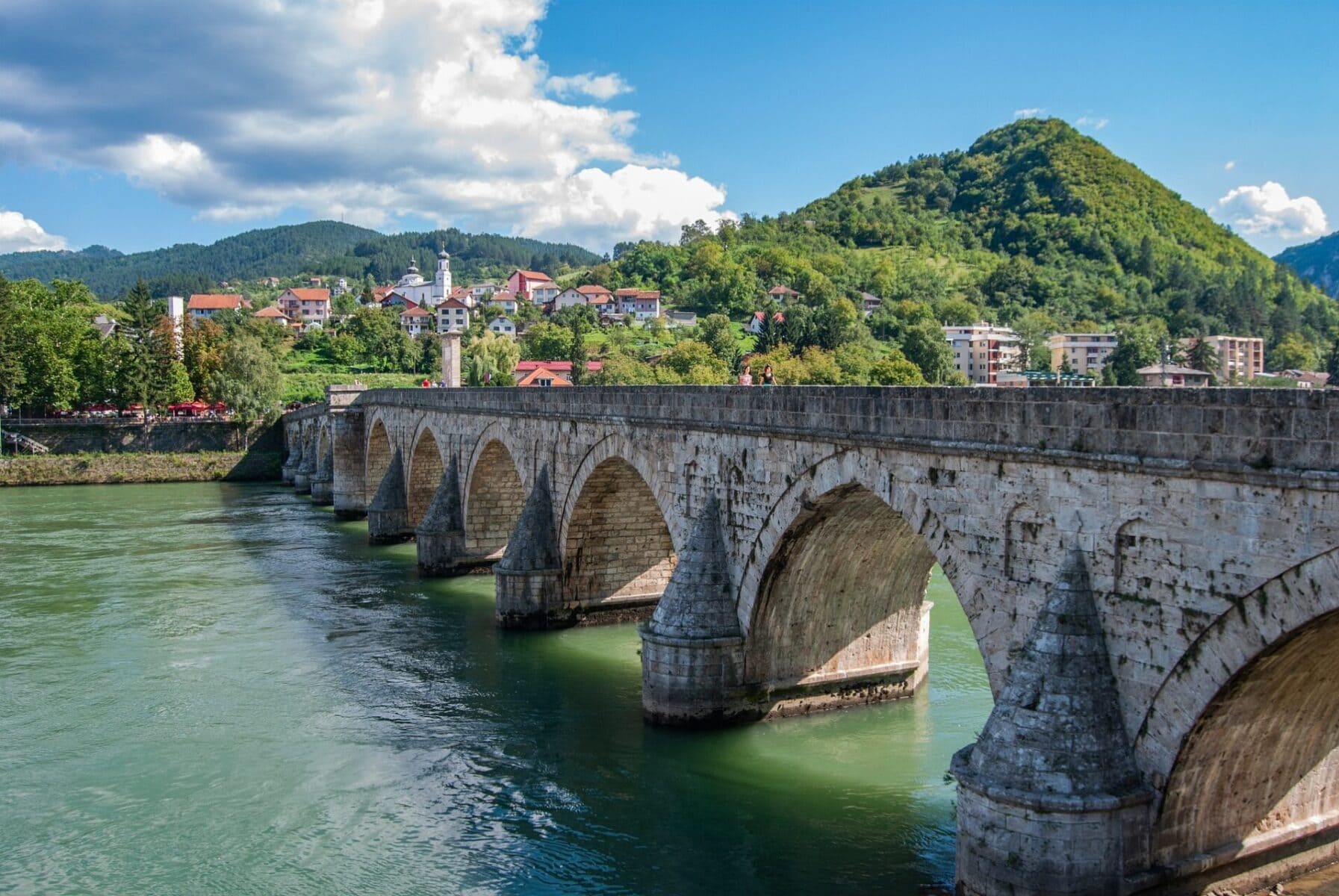 Historic stone bridge crossing a green river with hills and houses in the background.