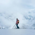 Skier standing on a snowy mountain ridge surrounded by clouds and alpine mountains
