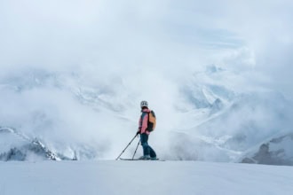 Skier standing on a snowy mountain ridge surrounded by clouds and alpine mountains