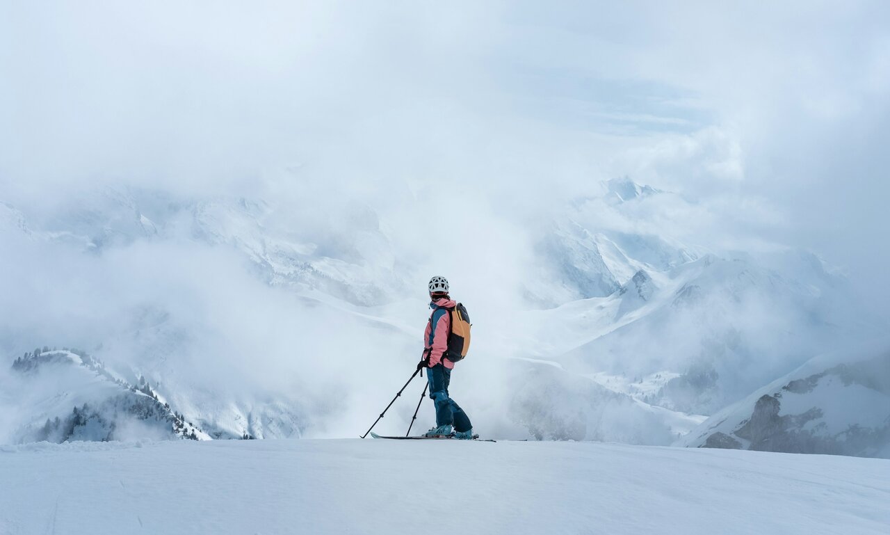 Skier standing on a snowy mountain ridge surrounded by clouds and alpine mountains