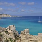 Turquoise coastal waters and limestone cliffs in the Egadi Islands near Favignana, Sicily, with a sailing boat anchored offshore.