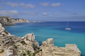 Turquoise coastal waters and limestone cliffs in the Egadi Islands near Favignana, Sicily, with a sailing boat anchored offshore.