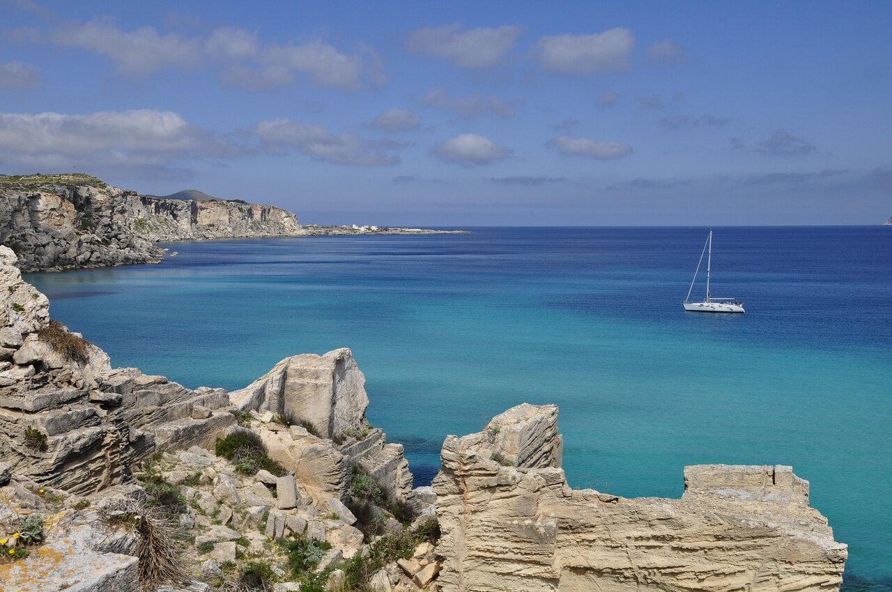 Turquoise coastal waters and limestone cliffs in the Egadi Islands near Favignana, Sicily, with a sailing boat anchored offshore.