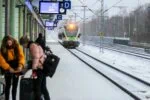 Passengers waiting beside a snowy commuter train platform in Finland as a green-and-white airport train arrives during winter.