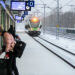 Passengers waiting beside a snowy commuter train platform in Finland as a green-and-white airport train arrives during winter.