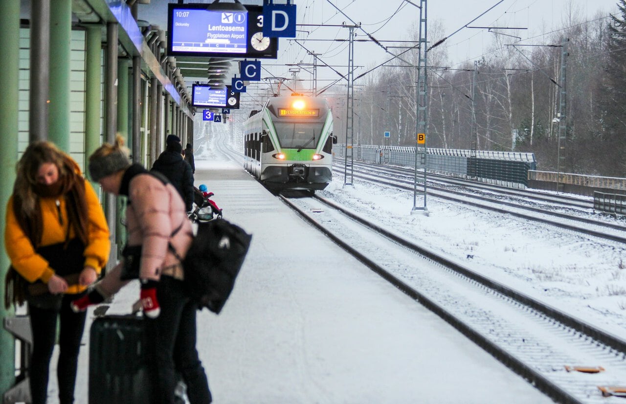 Passengers waiting beside a snowy commuter train platform in Finland as a green-and-white airport train arrives during winter.