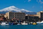 Snow-covered Mount Etna rising behind the harbour and fishing boats in Catania, Sicily, on a clear winter day.