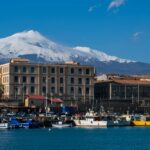Snow-covered Mount Etna rising behind the harbour and fishing boats in Catania, Sicily, on a clear winter day.