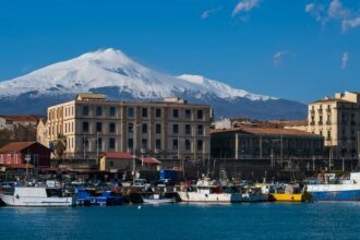 Snow-covered Mount Etna rising behind the harbour and fishing boats in Catania, Sicily, on a clear winter day.