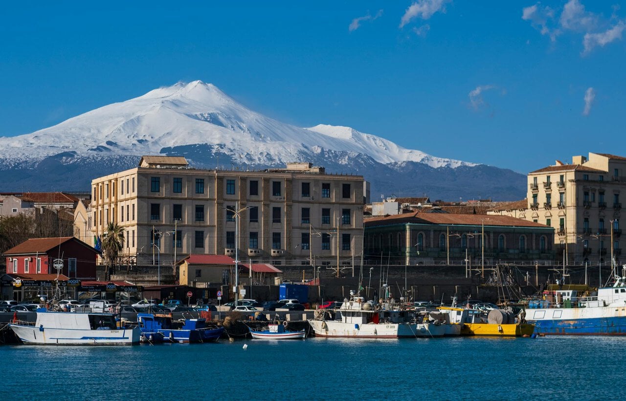 Snow-covered Mount Etna rising behind the harbour and fishing boats in Catania, Sicily, on a clear winter day.