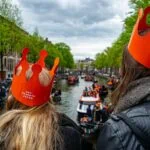 Travellers wearing orange crowns overlooking crowded Amsterdam canals during King’s Day celebrations in the Netherlands