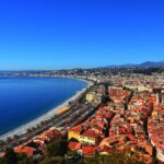 Panoramic view of Nice and the Promenade des Anglais on the French Riviera, showing the Mediterranean coastline, beaches, Old Town rooftops, and surrounding hills.