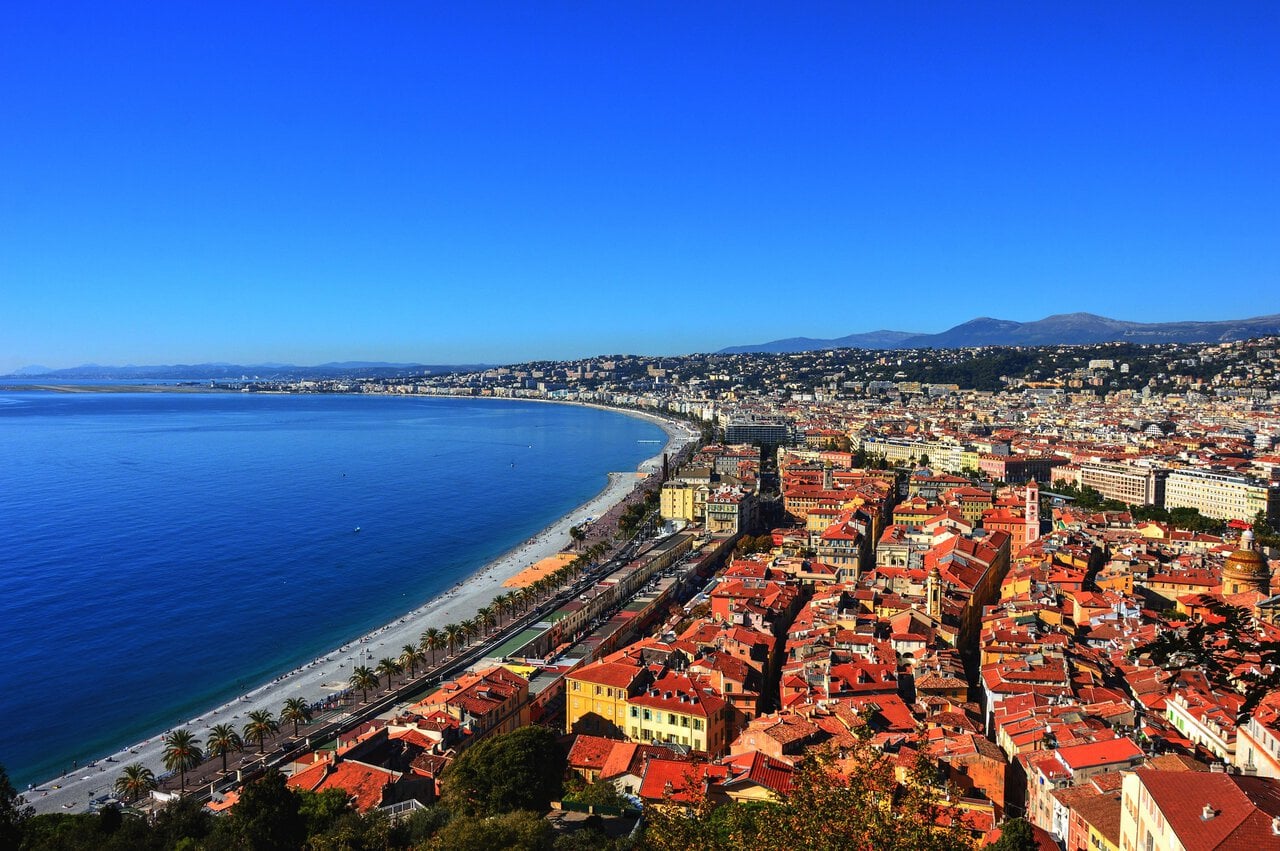 Panoramic view of Nice and the Promenade des Anglais on the French Riviera, showing the Mediterranean coastline, beaches, Old Town rooftops, and surrounding hills.