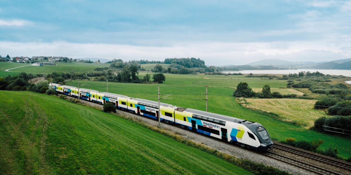 WESTbahn double-decker train travelling through the Austrian countryside near a lakeside landscape with rolling green fields and distant hills.