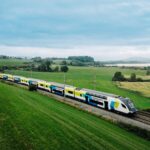 WESTbahn double-decker train travelling through the Austrian countryside near a lakeside landscape with rolling green fields and distant hills.