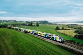 WESTbahn double-decker train travelling through the Austrian countryside near a lakeside landscape with rolling green fields and distant hills.