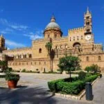 Palermo Cathedral in Sicily with its Norman towers, large central dome and historic square under a blue sky.