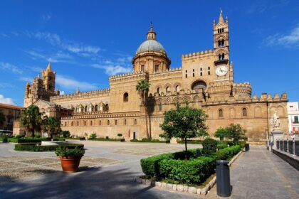 Palermo Cathedral in Sicily with its Norman towers, large central dome and historic square under a blue sky.