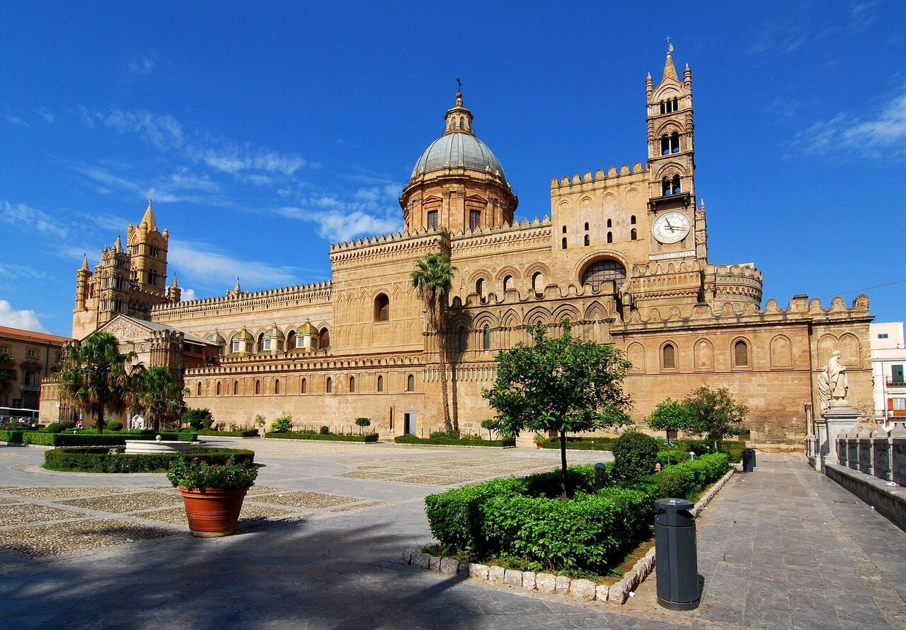 Palermo Cathedral in Sicily with its Norman towers, large central dome and historic square under a blue sky.